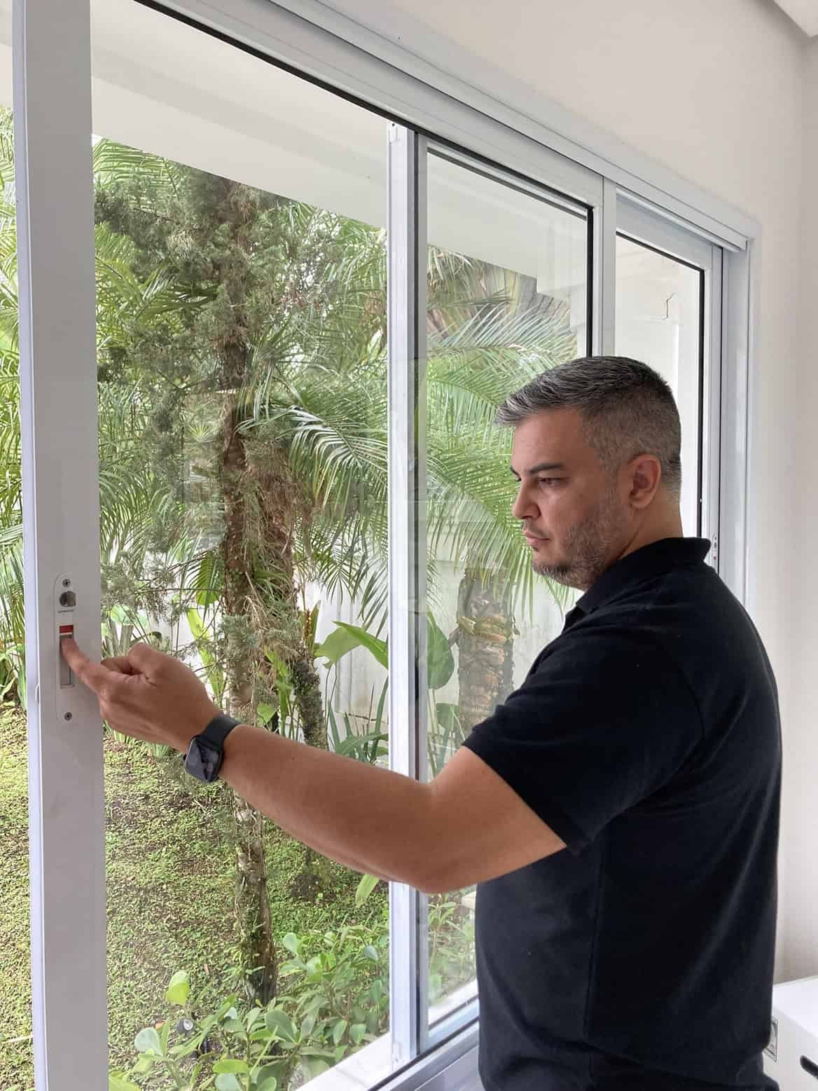 Janela de vidro com homem ajustando o puxador, vista para jardim com árvores verdes, iluminação natu.